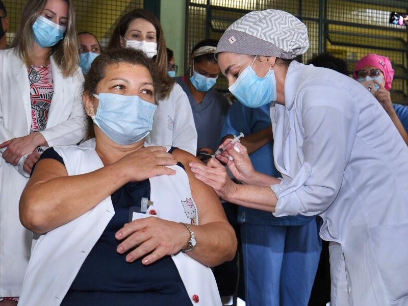 Uma enfermeira aplica a vacina no braço esquerdo de Maria Eliana, que olha para o lado. Atrás dela, algumas pessoas acompanham o momento
